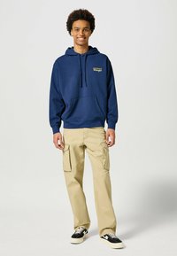 Granatowa bluza z kapturem i przednią kieszenią, żółte logo. Beżowe spodnie cargo z bocznymi kieszeniami, połączone z czarno-białymi trampkami.