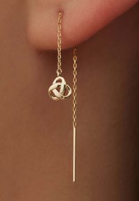 Boucles d'oreilles en chaîne dorée avec un pendentif en forme de nœud et un accent de tige droite et élancée. Fini brillant avec des détails complexes sur le pendentif.