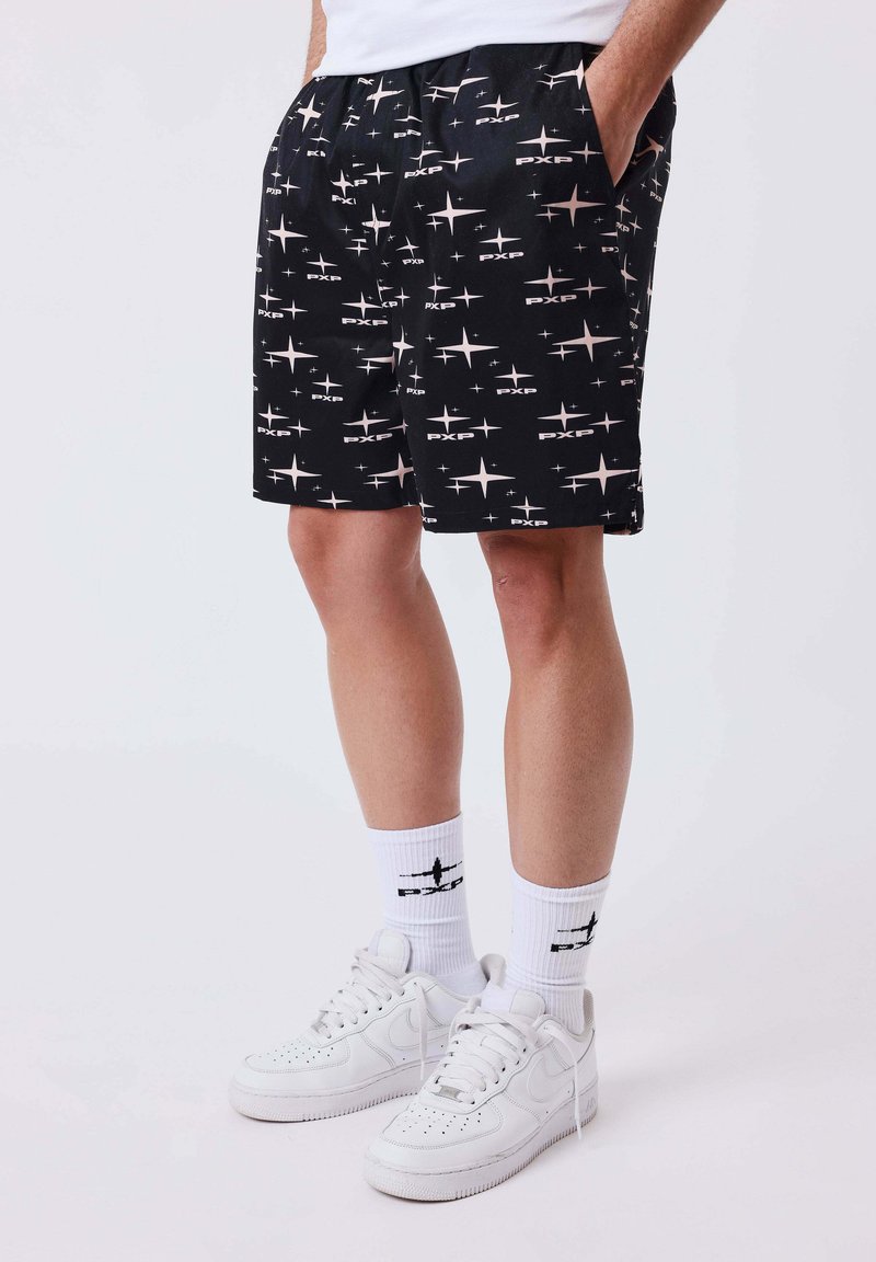 Personne portant un short noir avec des motifs d'étoiles blanches, des chaussettes blanches avec un motif noir, et des baskets blanches de sport, debout les mains dans les poches.