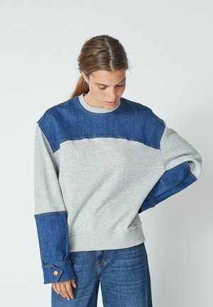 Gray sweatshirt with blue denim accents on shoulders and sleeves. Features a rounded neck and button details on cuffs. Casual fit.