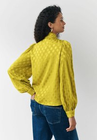 Senfgelbe Bluse aus strukturiertem Stoff, mit puffigem Schnitt, Knopfmanschetten und kreisförmigen Mustern. Kombiniert mit blauen Jeans.