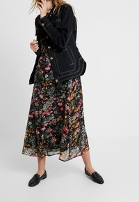 Veste en denim noire avec des coutures blanches, portée sur une robe maxi fleurie avec des motifs roses, oranges et blancs sur un fond sombre. Chaussures plates noires.