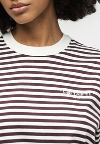 Κοντινό πλάνο ενός ατόμου που φοράει ένα ριγωτό πουκάμισο Carhartt WIP σε λευκό και σκούρο καφέ, με λευκή ριμπ απολήξη στο γιακά.