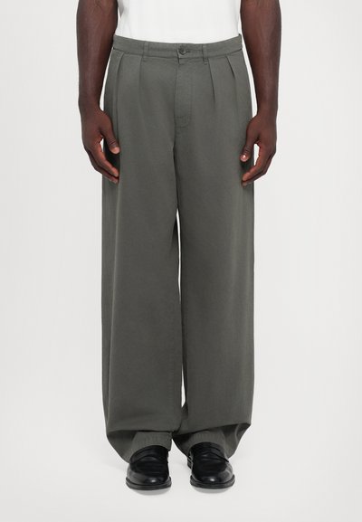 Pantalons à jambe large en tissu vert foncé, dotés de plis sur le devant, d'une fermeture à bouton et de poches latérales. Portés avec des chaussures noires brillantes.