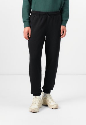 Black sweatpants with an elastic waistband and cuffs, featuring a drawstring detail. Worn with cream-colored athletic shoes.