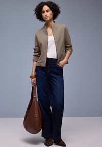 Femme aux cheveux bouclés portant une veste taupe, un t-shirt blanc, un jean large foncé, des bottes marron, tenant un grand sac en cuir marron, debout devant un fond gris.