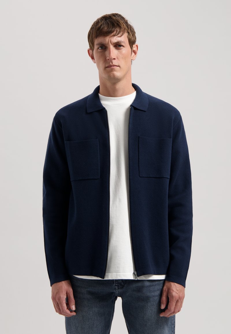Marineblauwe zip-up jas van gestructureerde stof, met twee voorste opgestikte zakken, een kraag en een ontspannen pasvorm over een wit shirt.