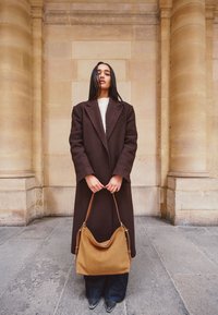 Brown wool coat over white sweater, dark wide-leg jeans, holding a tan leather bag with curved shape and long strap, set against a stone wall.