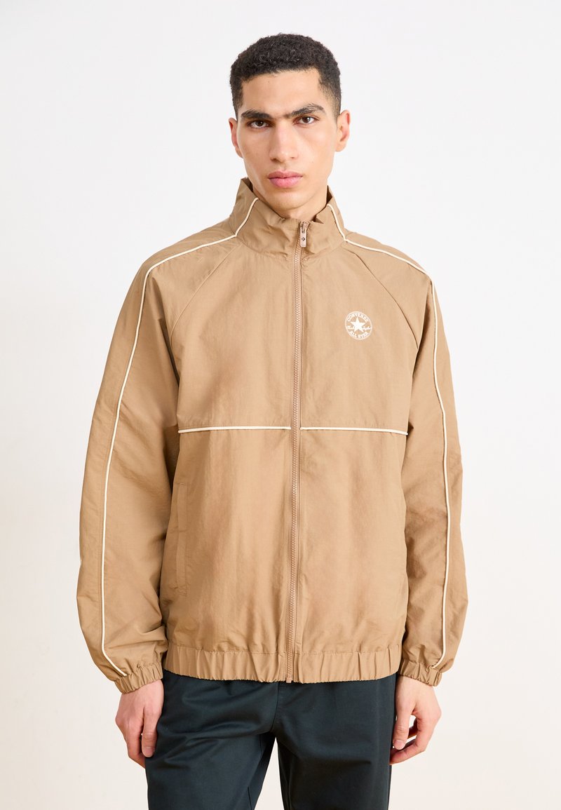 Veste coupe-vent marron avec un col haut, des accents blancs, des poignets et une taille élastiques. Présente un logo sur le côté supérieur gauche de la poitrine.