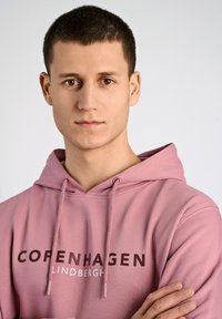 Sudadera rosa con cordón, que presenta "COPENHAGUE" en letras oscuras y en negrita, y "LINDBERGH" en una fuente más clara. Textura suave.