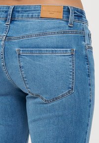 Blå denimjeans med en tettsittende passform, to baklommer og et brunt lærmerke i livet med preget tekst.
