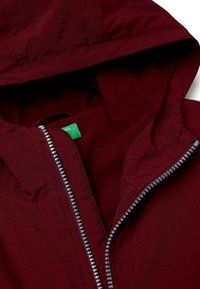 Giacca bordeaux con cappuccio, dotata di cerniera grigia, morbido rivestimento interno in pile e etichetta verde all'interno. Tessuto esterno resistente e testurizzato.