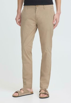 Pantaloni beige a taglio dritto indossati con sandali marrone chiaro in suede a due fasce, su una persona in piedi davanti a uno sfondo uniforme.