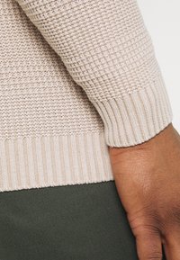 Beżowy sweter o fakturze z prążkowanymi mankietami, z wzorem dzianiny. Tło przedstawia ciemnozielony materiał, co sugeruje stylizację warstwową.