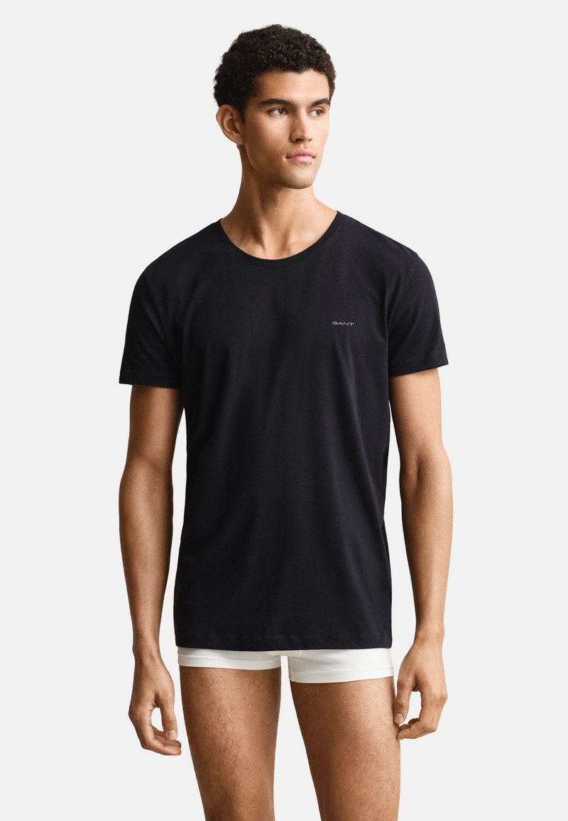 Mann trägt ein schlichtes schwarzes T-Shirt mit kurzen Ärmeln und weiße Boxershorts, schaut leicht zur Seite vor schlichtem Hintergrund.