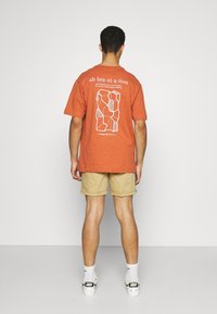 Rost-orange Baumwoll-T-Shirt mit weißem Grafikdesign auf der Rückseite, das abstrakte Formen und Text zeigt. Eine beige Shorts und weiße Sneaker.
