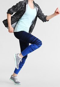 Idrottskläder bestående av en ljusblå linne, mönstrade blå leggings och grå sportskor med gröna detaljer, i lager med en svart jacka.