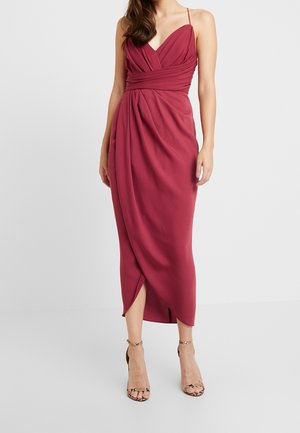 Femme portant une robe portefeuille sans manches rouge profond avec un décolleté en V et de fines bretelles, associée à des sandales à talons hauts avec bride à la cheville.