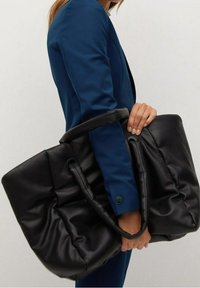 Sac à main noir rembourré avec deux poignées robustes, en matériau lisse, présentant une texture douce et un design minimaliste.