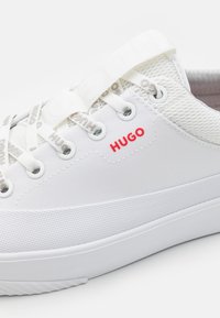 Bílé plátěné tenisky s průsvitnými akcenty, plochými tkaničkami a texturovanou gumovou podrážkou. Na boku se nachází červené logo "HUGO".