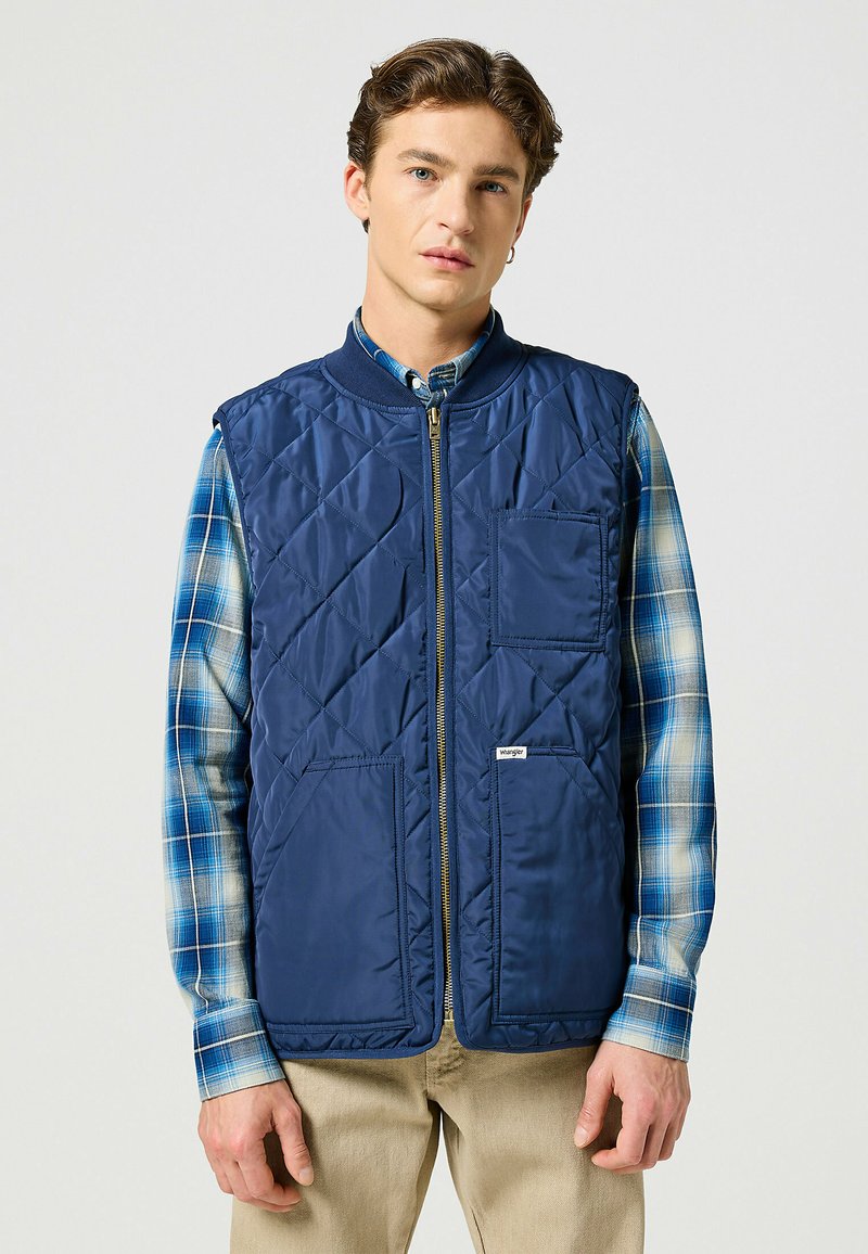 Gilet trapuntato blu navy con colletto alto, zip frontale e tasca a toppa; indossato sopra una camicia a quadri blu e bianca, con una vestibilità strutturata.