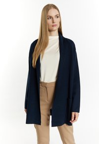 Cardigan aperto blu navy in tessuto morbido, con maniche lunghe, tasche laterali e vestibilità comoda. Abbinato a una maglia crema e pantaloni beige.