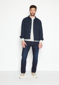 Marineblauwe denimjas met drukknoopjes, twee borstzakken; lichtgrijze trui eronder; donkere spijkerbroek; witte sneakers met beige accenten.