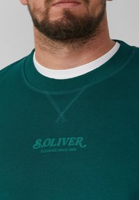 Felpa verde scuro con scollo rotondo e bordi a costine. Presenta una stampa logo sottile al centro e un colletto interno bianco a contrasto.