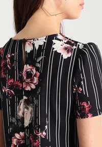 Svart tyg med blommönster som har vertikala vita ränder. Rund halsringning med nyckelhålsdetalj i ryggen och korta puffärmar.