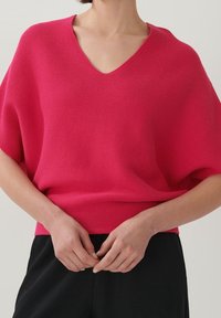 Fuchsiový svetr s V-výstřihem, pletený, s širokými rukávy, ribbovanou texturou a volným střihem, v kombinaci s černými kalhotami.