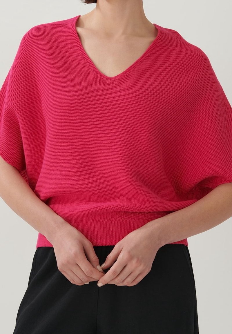 Fuchsiový svetr s V-výstřihem, pletený, s širokými rukávy, ribbovanou texturou a volným střihem, v kombinaci s černými kalhotami.