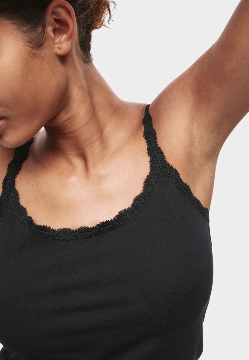 Zwarte tanktop met geschulpte kanten rand langs de halslijn en schouderbanden. Zacht, rekbaar materiaal benadrukt een getailleerde vorm.