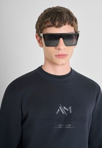 Gafas de sol negras con diseño angular y lentes oscuros, combinadas con una sudadera azul marino con un logo en relieve y cuello redondo.