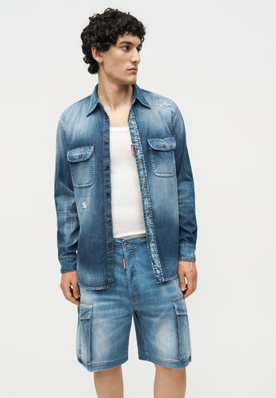 Νέος άνδρας που φοράει ξεθωριασμένο μπλε τζιν πουκάμισο πάνω από λευκό αμάνικο και ταιριαστά τζιν cargo σορτς, κοιτάζοντας προς τα πλάγια.