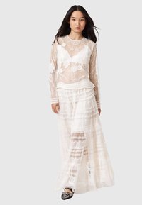 Λευκό στρωματοποιημένο δαντελένιο φόρεμα με μακριά διαφανή μανίκια, κεντητά λουλουδάτα σχέδια και κυματιστά διακοσμητικά στοιχεία. Ημι-διάφανη δαντέλα πάνω από σταθερό υπόστρωμα.