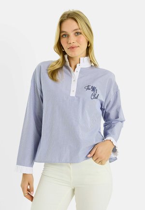 Mujer joven con pantalones blancos y una camisa de manga larga azul claro con rayas finas y el texto "The 90's Club" en el pecho, posando con una mano en el bolsillo.