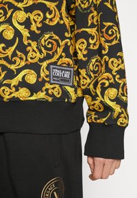 Sudadera negra con un intrincado patrón de espirales doradas, que presenta un puño y dobladillo acanalados. Incluye una etiqueta negra con texto blanco de Versace Jeans Couture.