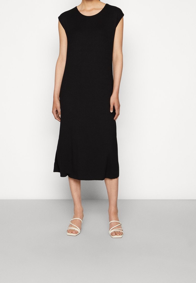 Femme portant une robe midi noire sans manches et des sandales blanches à talons avec brides, debout devant un fond uni.