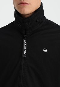 Czarna kurtka z wysokim kołnierzem, na zamek błyskawiczny i napy. Posiada logo z tkaniny oraz fakturowany materiał. Dopasowany krój.