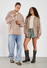 Two models wear casual outfits. The male model has a beige bomber jacket, light jeans, and sneakers. The female model wears a beige jacket, light t-shirt, denim shorts, and knee-high black boots.