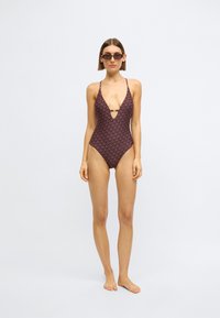 Maillot de bain une pièce violet foncé à motif géométrique, doté d'un décolleté plongeant et de bretelles fines, offrant une silhouette ajustée.