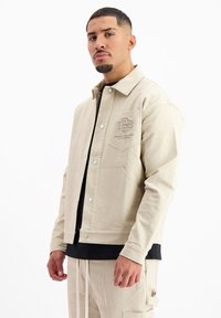 Beige corduroy jas met een kraag, voorzakken en een geborduurd logo op de borst. Glad textuur, knoopsluiting.