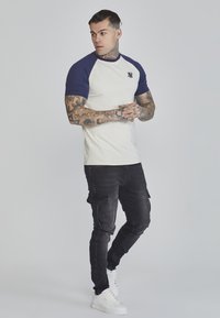 Raglan-T-Shirt, cremfarbener Körper mit marineblauen Ärmeln, Logo auf der Brust. Kombiniert mit schwarzen Cargo-Hosen und weißen Sneakern, die Tattoos an den Armen zur Schau stellen.