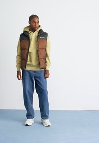 Man in een bruin en zwart gewatteerd vest over een beige hoodie, met losse blauwe jeans en witte sneakers, die tegen een witte muur en blauwe vloer staat.
