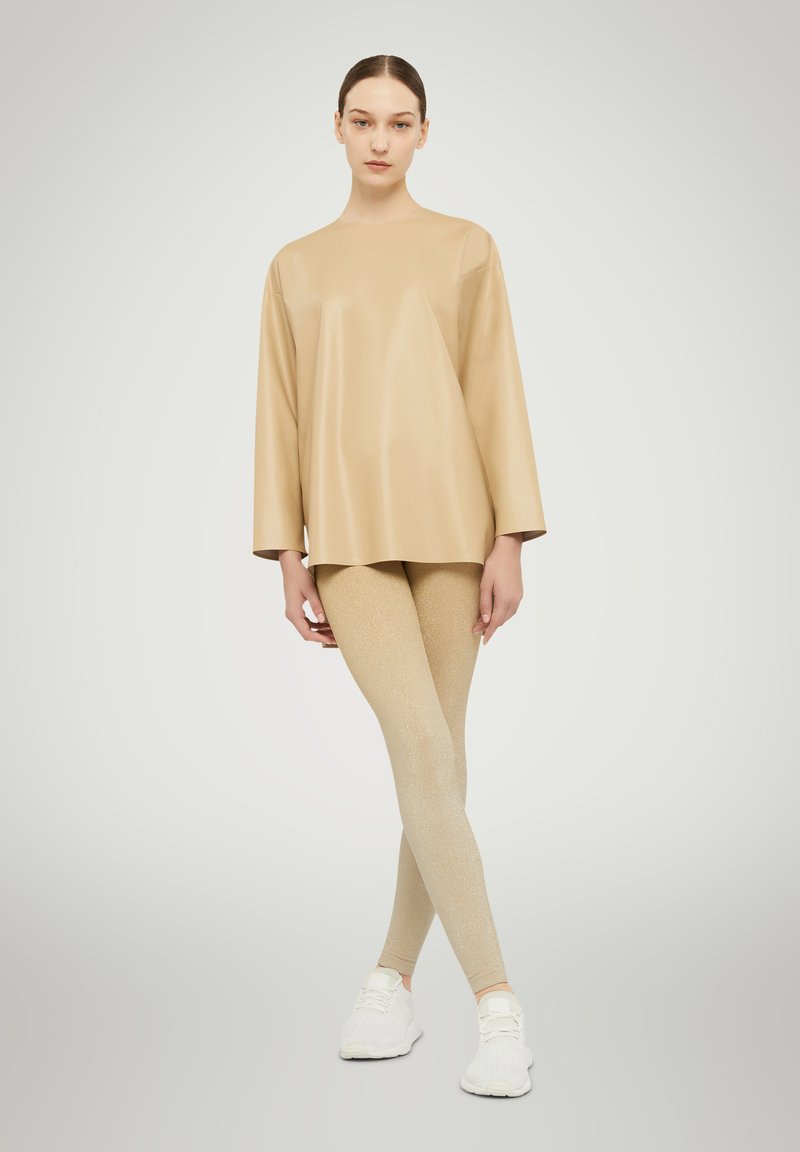 Beige synthetisches Oberteil mit Dreiviertelärmeln, kombiniert mit hellgoldenen, glitzernden Leggings und weißen Sportschuhen. Glatte Texturen, minimales Design.