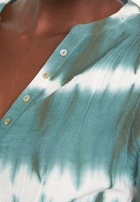 Camicia abbottonata in tessuto leggero con motivo tie-dye in turchese e bianco, dotata di colletto e tre bottoni sullo scollo.