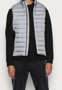 Helles graues gepolstertes Weste mit horizontalem Steppmuster, Reißverschluss und schwarzen Akzenten, getragen über einem schwarzen Langarmshirt.