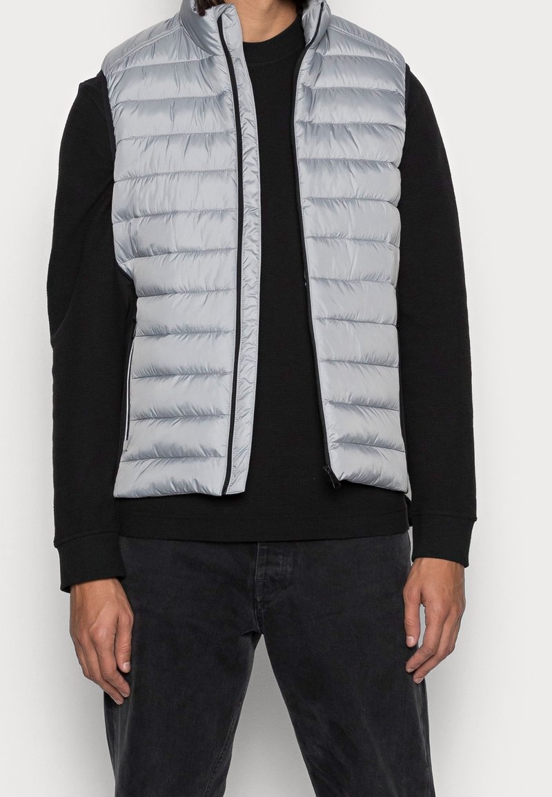 Helles graues gepolstertes Weste mit horizontalem Steppmuster, Reißverschluss und schwarzen Akzenten, getragen über einem schwarzen Langarmshirt.