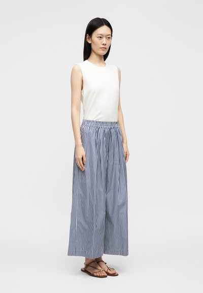 Femme debout portant un haut blanc sans manches, un pantalon large à rayures verticales bleues et blanches, et des sandales plates marron avec des brides à la cheville.