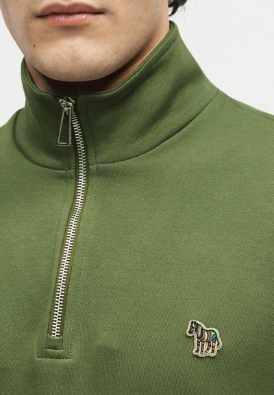 Κοντινό πλάνο ενός ατόμου που φοράει πλεκτό πουλόβερ ελιάς με φερμουάρ στο τέταρτο μέρος και πολύχρωμο ριγέ έμβλημα αλόγου στο στήθος.